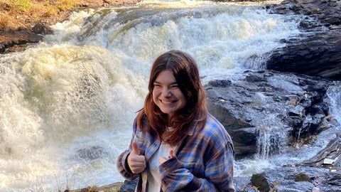 A smiling young woman wearing a plaid shirt stands in front of a mountain stream
