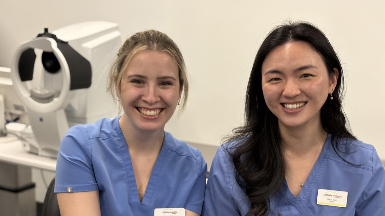 Two young women wearing blue scrubs and name tags are seated in a clinic with medical equipment visible in the background