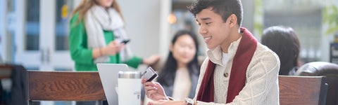 Student holds a credit card in one hand while working on their laptop