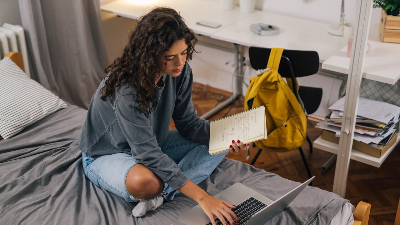 A young woman sits cross-legged on a bed with grey bedding, using a laptop