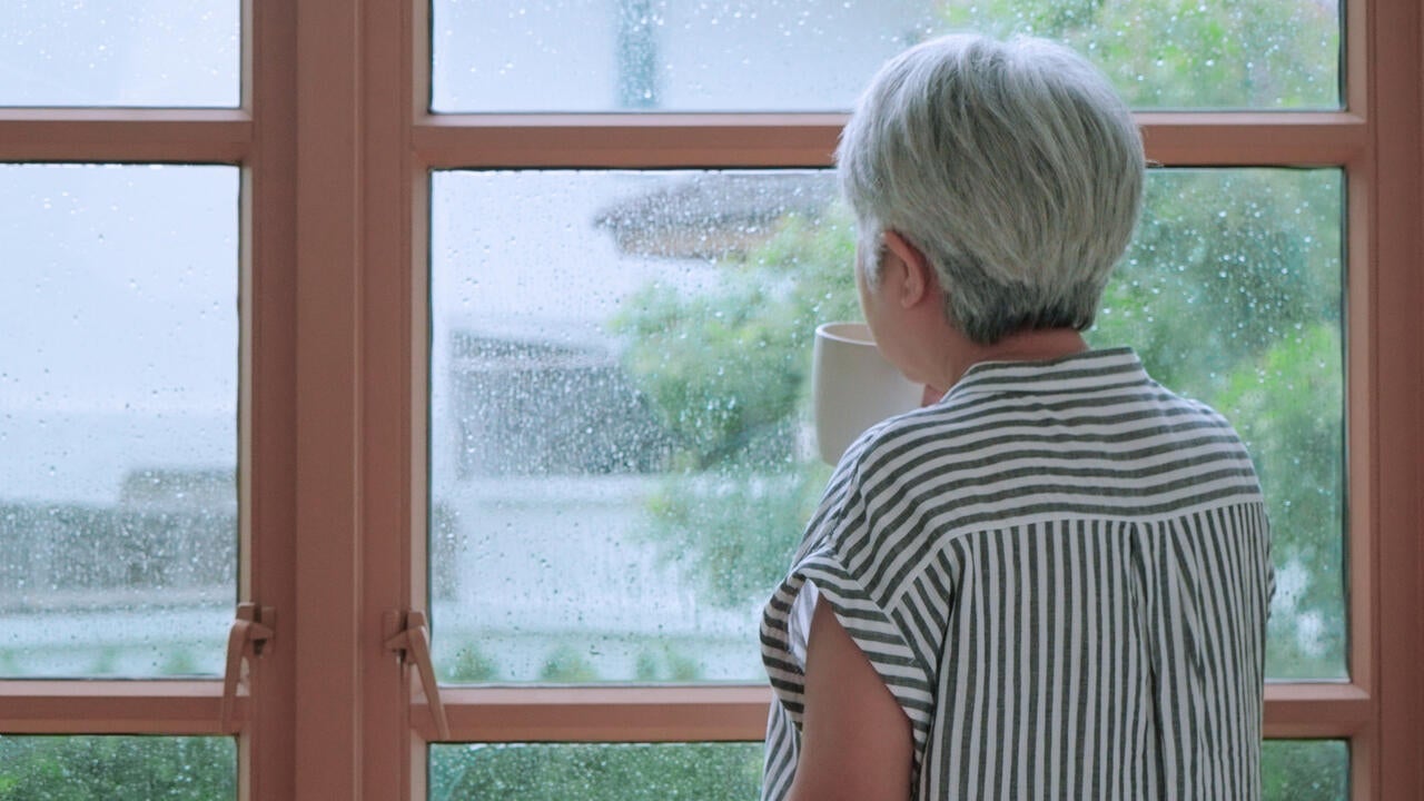 A senior person at home drinking from a cup while looking out a window.