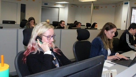 A woman with blonde hair and glasses is speaking on the phone, while another woman with brown hair types on a laptop