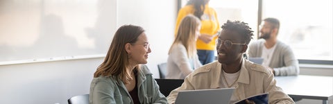 Two students talking at table with other students in background