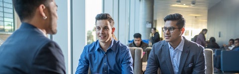 Three business students in suits sitting next to brightly lit window, engaged in professional discussion while smiling.