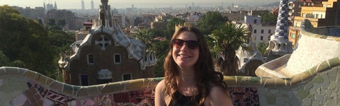 A woman wearing sunglasses smiles while sitting on a mosaic bench with a european cityscape in the background.