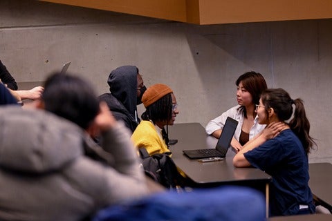 Group of four students in a lecture hall sitting at a table together around a laptop