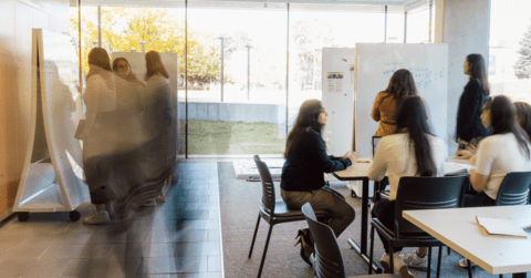 Many students in a room together - some gathered at a whiteboard and some at a table, image is blurred for an in-motion or action effect 
