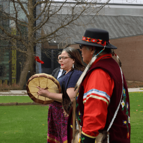 Two people in traditional clothing take part in a cultural ceremony, one holding a hand drum outdoor
